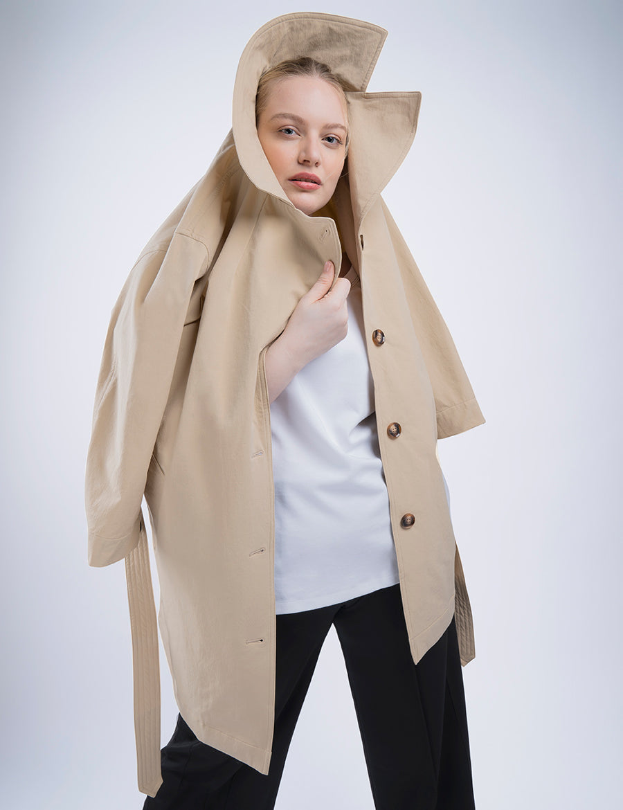 Plus size model wearing loose navy blue pants and a loose white shirt. She wrapped a beige trench coat with three buttons around her shoulders and with one hand is holding the top part over her head like a hood.